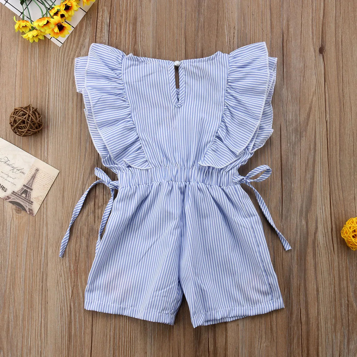 Blue and white striped toddler romper with ruffled sleeves and tie detailing on a wooden surface, surrounded by a Paris-themed postcard, yellow flowers, and decorative balls.