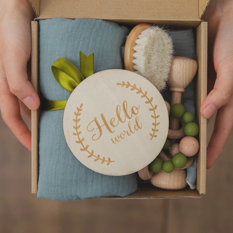 Hands holding a box containing a baby gift set with a wooden "Hello world" plaque, blue cloth, green beaded teething toy, soft brush, and wooden utensils tied with a green ribbon.