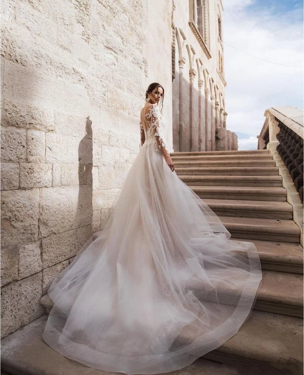 Bride in a lace wedding gown with a flowing train stands on stone steps beside an ancient building, glancing back. The sky is clear with a few clouds in the background.