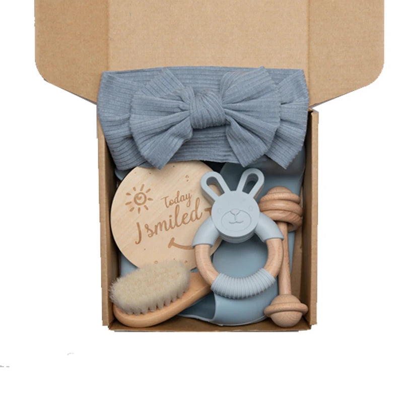 A brown box containing a gray headband, a wooden disc with "Today I Smiled," a wooden-handled brush, a blue-gray rabbit teether, and a knotted rope toy.