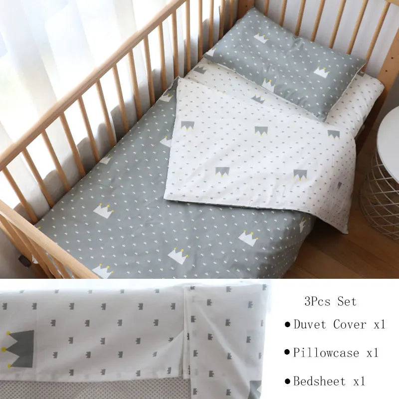 Gray and white three-piece crib bedding set with a crown pattern, including a duvet cover, pillowcase, and bedsheet, displayed in a wooden crib next to a small round table.