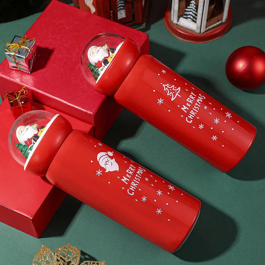 Red Christmas-themed thermoses with snow globe lids featuring Santa, surrounded by red gifts and ornaments on a green surface. Each thermos says "Merry Christmas" with snowflakes.