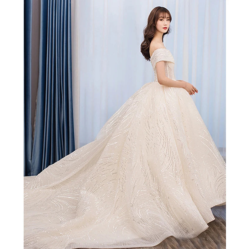 Woman in an elegant off-shoulder ivory wedding gown with a flowing, embellished train stands in profile against a backdrop of blue curtains, showcasing classic bridal style.