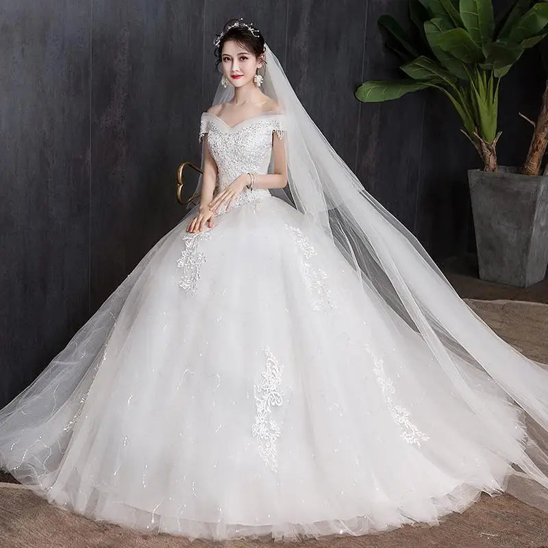 Bride in an elegant off-shoulder, lace-embellished white gown with a long veil, standing against a dark background beside a large potted plant.