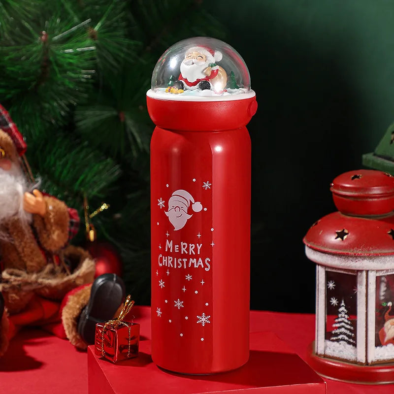 Red Christmas-themed bottle with Santa figure inside a snow globe on top. “Merry Christmas” text and snowflakes on the side. Surrounded by holiday decorations.