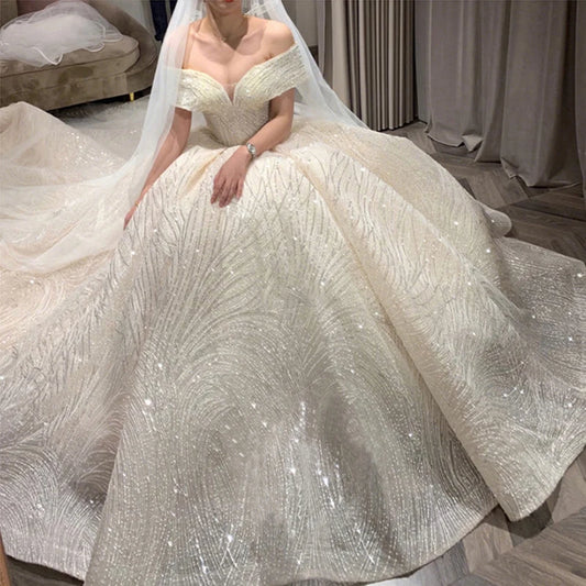 Bride in an off-shoulder, sparkling ivory ball gown with a beaded pattern, seated gracefully. A long, sheer veil cascades behind her. Gown details glisten under soft lighting.