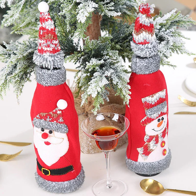 Festive bottle covers resembling Santa and a snowman with red outfits and winter hats, placed beside a Christmas tree, a glass of amber liquid, and gold cutlery.