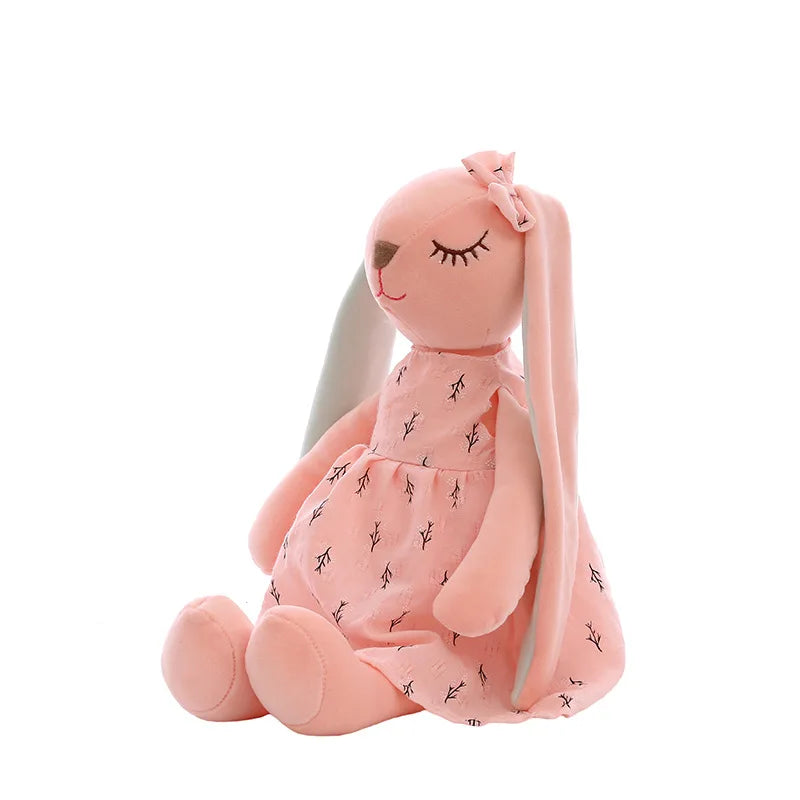 A plush pink bunny with closed eyes, wearing a matching dress with a delicate branch pattern and a bow on its head. The toy is seated against a white background.