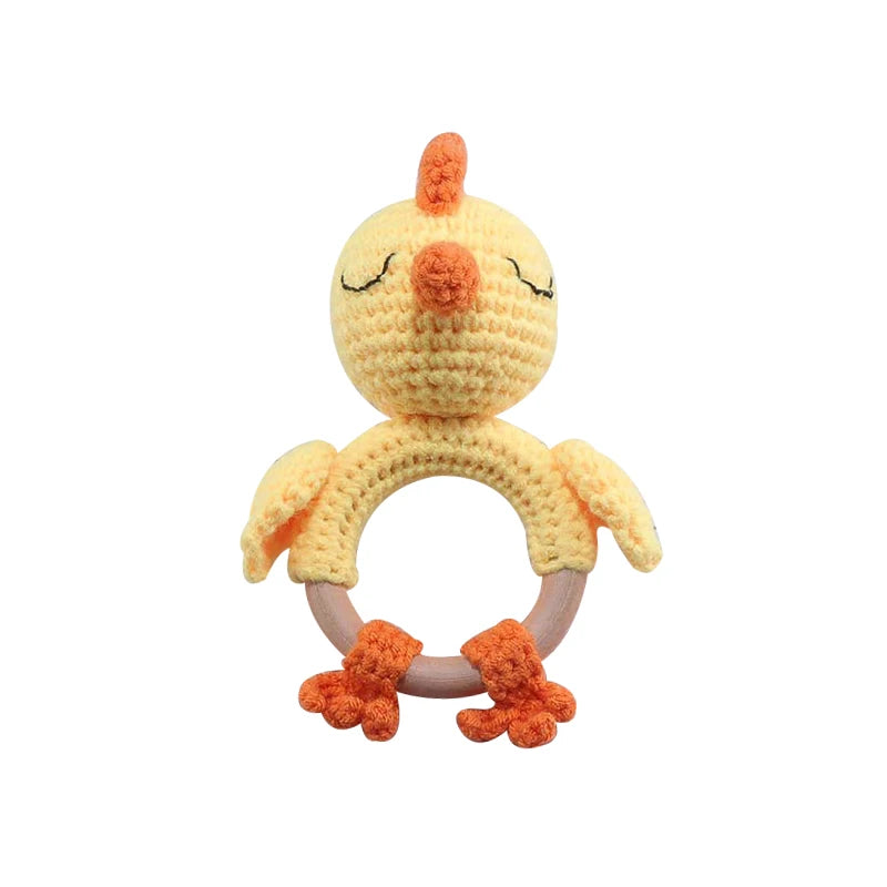 Crocheted chick-shaped baby rattle with a circular wooden ring. The chick has orange feet and beak, closed eyes, and light yellow body, resembling a cute, sleepy bird.
