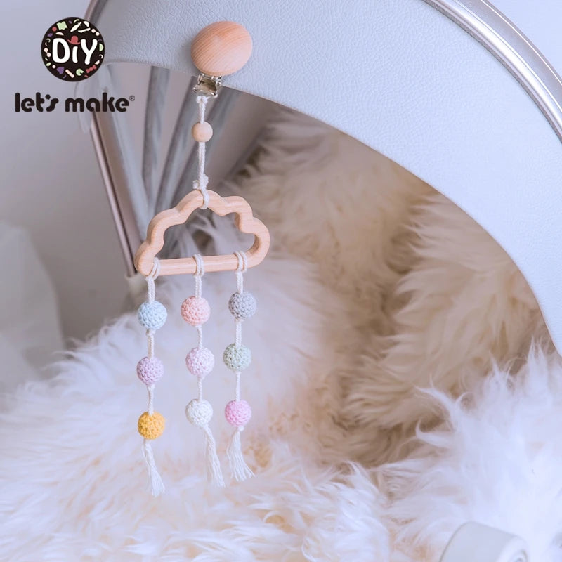Wooden cloud-shaped baby mobile with pastel crochet beads hangs on a stroller with white fur lining. A logo on the top left reads "DIY let's make."