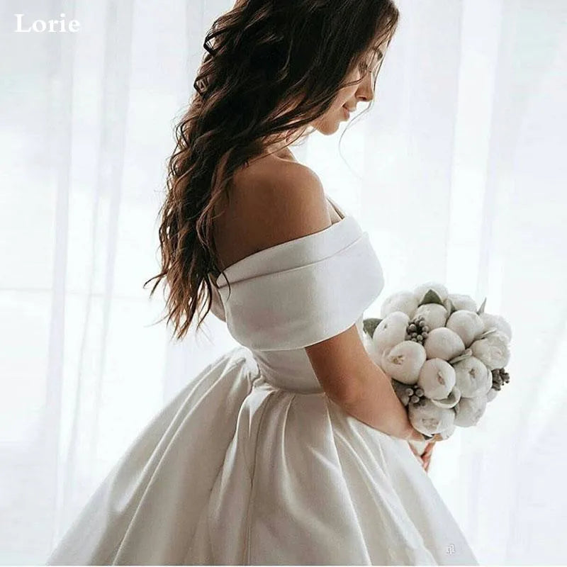 Bride in an off-shoulder white gown holding a bouquet of white flowers, standing by a sheer curtain with long, wavy hair cascading down her back.