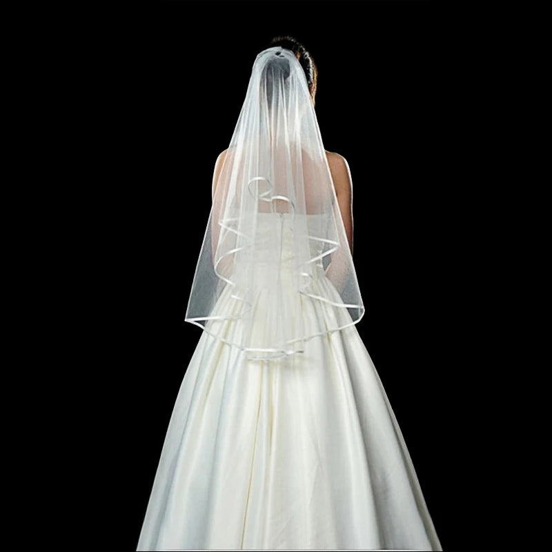 Bride in a white gown with a layered veil, viewed from behind against a black background. The dress has a full skirt, and the veil extends to the waist.