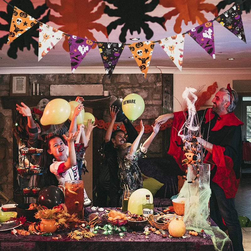 A festive Halloween party scene with people dressed in costumes, joyfully tossing balloons. The room is adorned with colorful spider and pumpkin bunting. A table in the foreground is decorated with themed items like a "toxic" bottle, cobwebs, and autumn leaves, enhancing the spooky atmosphere.
