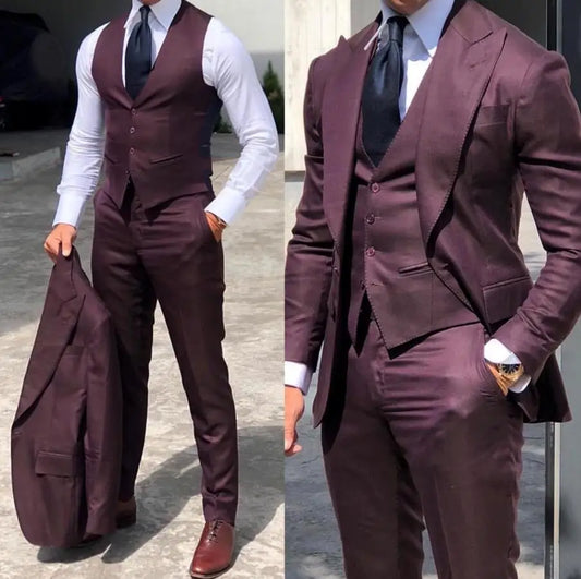Man wearing a burgundy three-piece suit with a white shirt and black tie. He holds the jacket in one hand, showcasing the vest and matching trousers. Brown shoes complete the look.