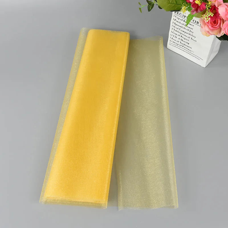 Yellow and translucent shimmer organza fabric sheets are layered on a gray surface beside a white paper bag with pink and green flower bouquet.
