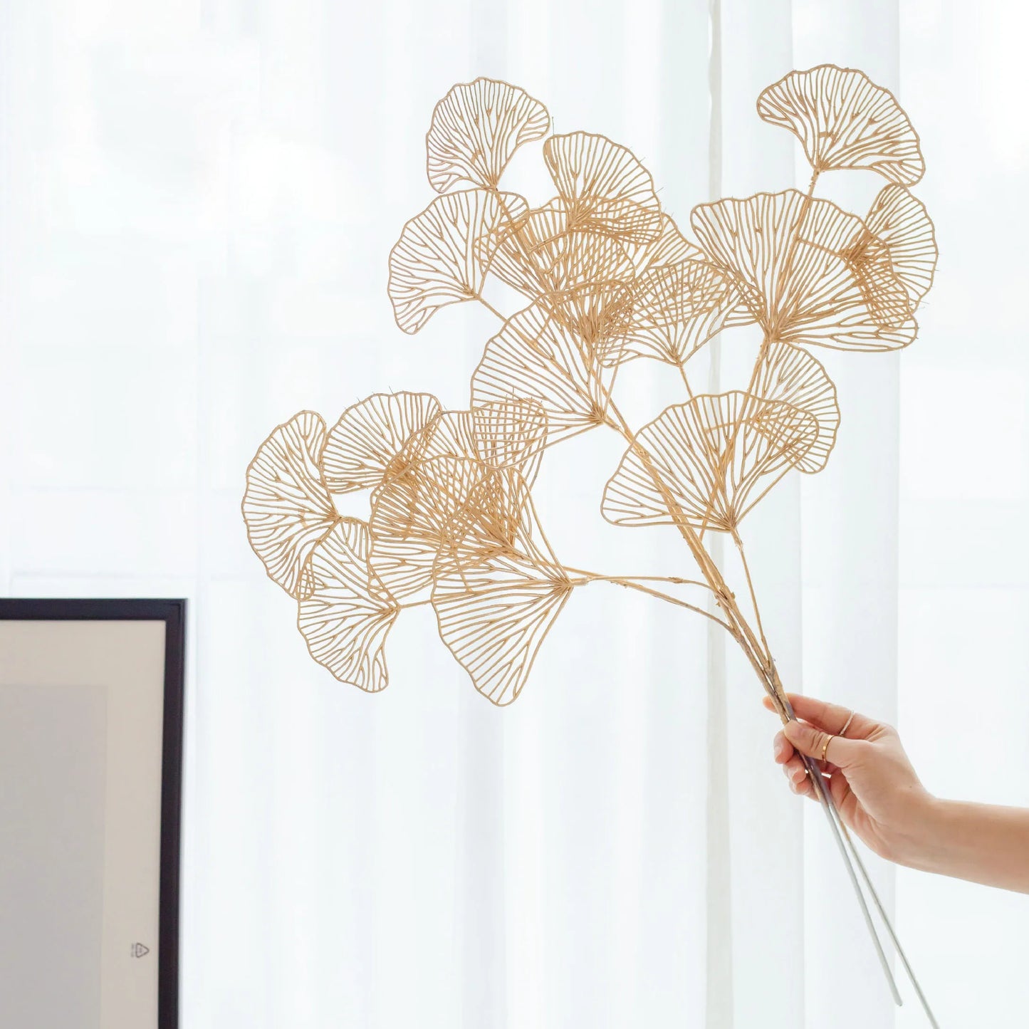 A hand holds delicate gold leaf-shaped wire branches against a softly lit white background, creating a minimalist and elegant display.