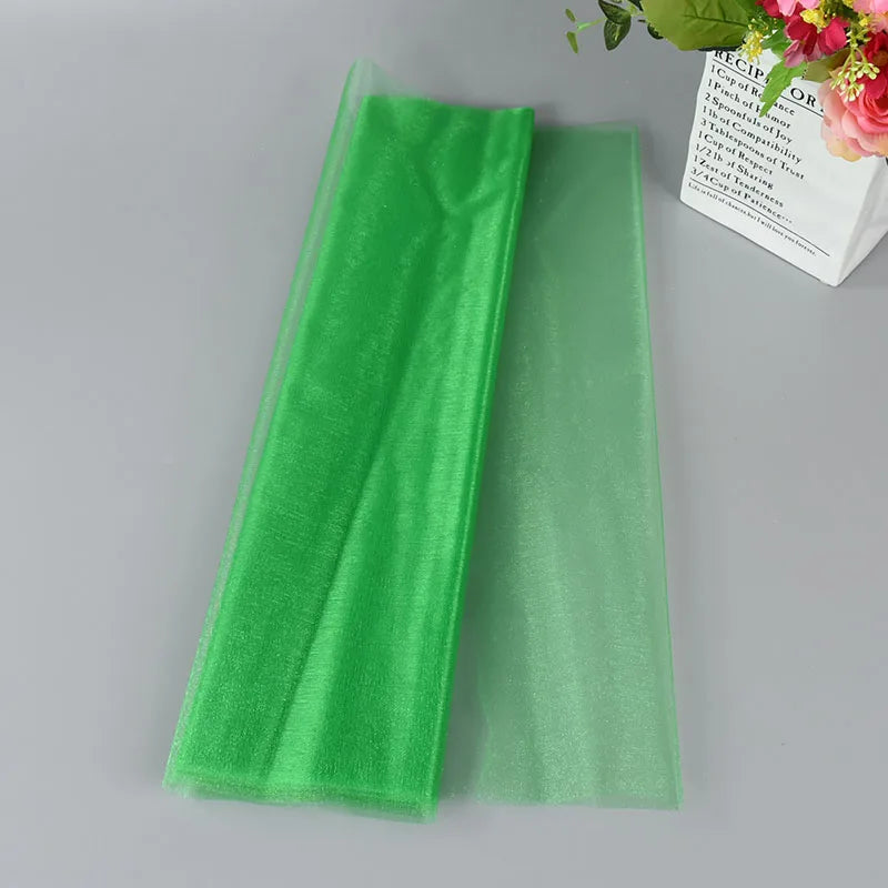 Two sheets of translucent green organza fabric are laid out on a gray surface, with a white paper bag containing pink flowers in the corner.