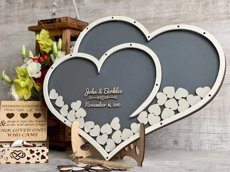 Two intertwined heart-shaped frames filled with small wooden hearts, marked with names and a date. Background includes flowers and a wooden guestbook box.