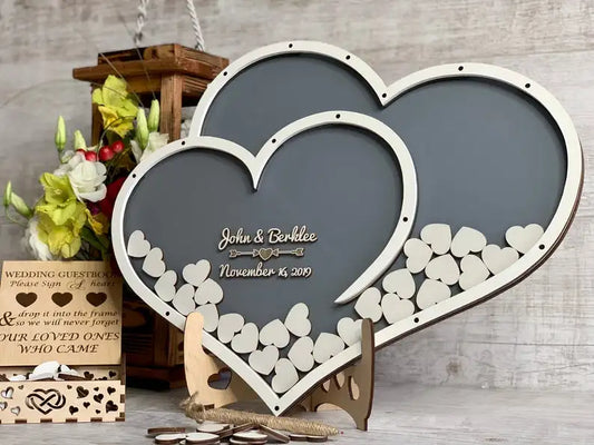 Two intertwined heart-shaped frames filled with small wooden hearts, marked with names and a date. Background includes flowers and a wooden guestbook box.