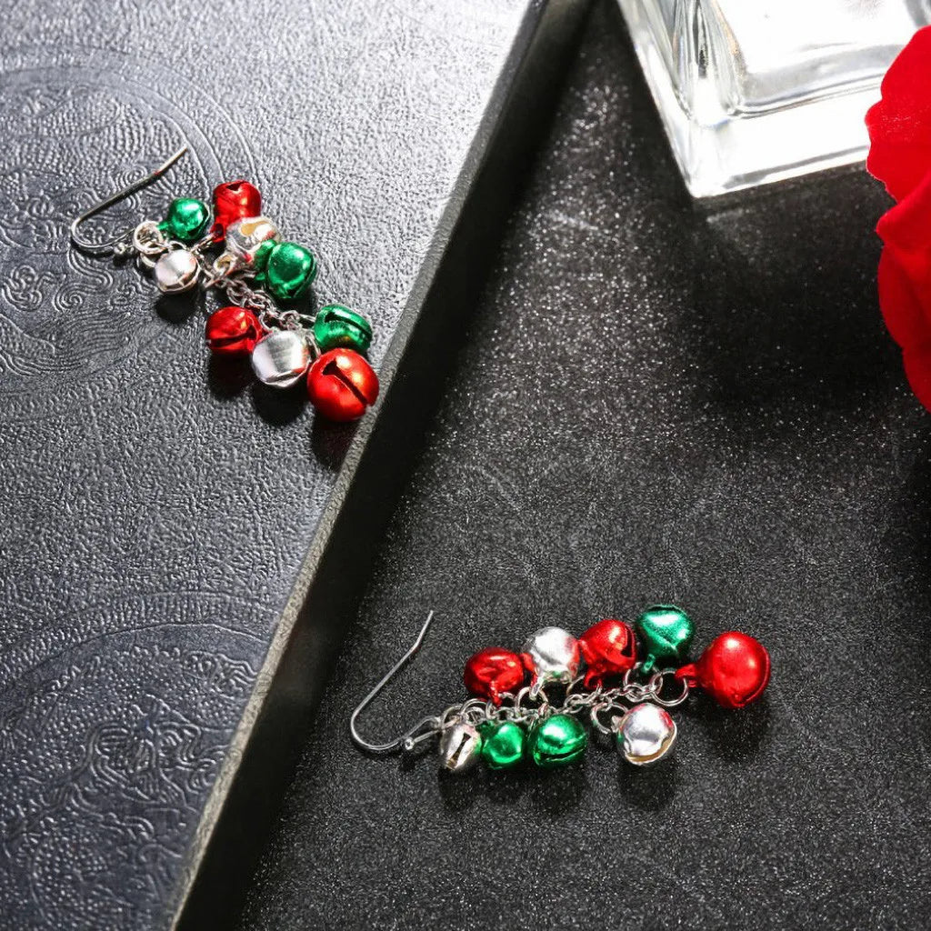 Festive earrings adorned with red, green, and silver jingle bells on a textured black surface. Adjacent is a red rose and glass bottle, creating a sophisticated holiday ambiance.