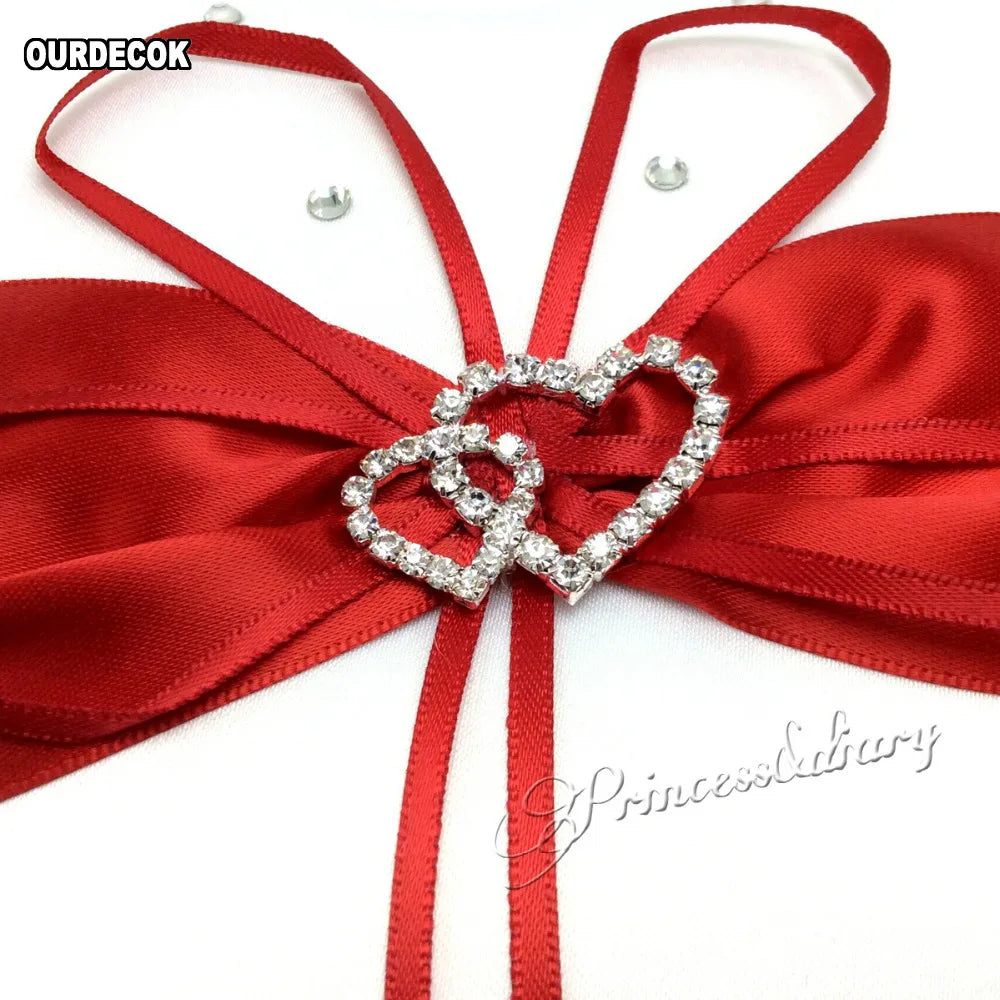 Red satin ribbon shaped into a bow with rhinestone-studded interlocking heart embellishment on white fabric. The words "PrincessDiary" and "OURDECOK" are visible.