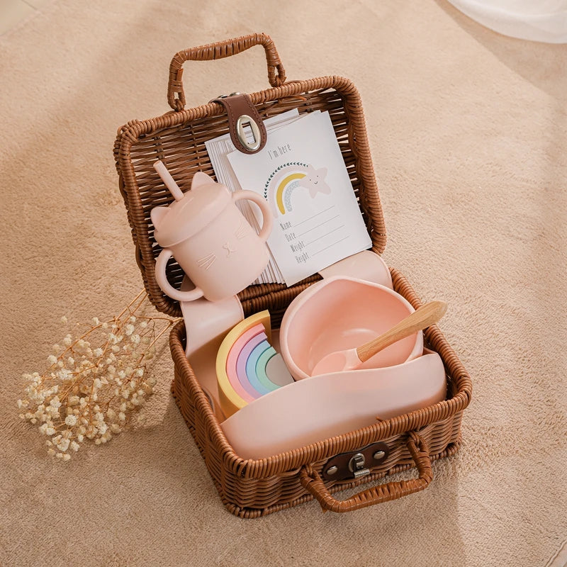 Wicker basket containing pastel-colored baby items: a pink sippy cup with ears, bowl and spoon, stacking rainbow toy, and cards with rainbow designs, on a soft beige surface.