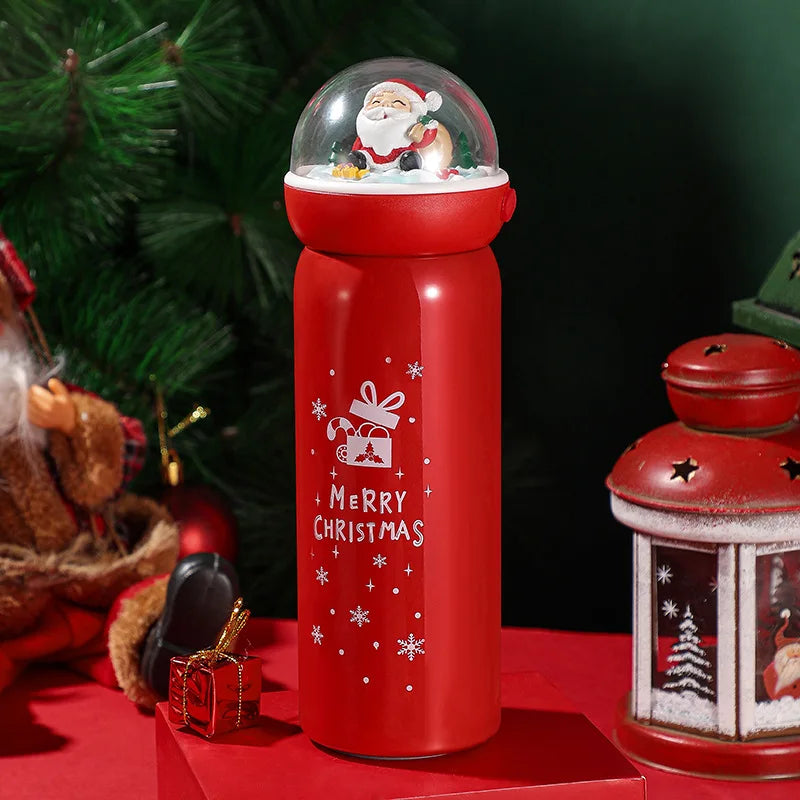Red Christmas-themed thermos with a Santa figurine globe on top. "Merry Christmas" text and gift illustration on the side. Background features holiday decorations and greenery.