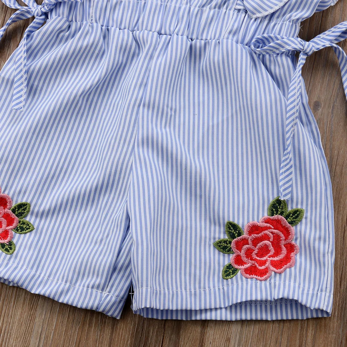 Blue and white striped shorts on a wooden surface, featuring red embroidered roses with green leaves at the hem. The shorts have a gathered waistband and tied sides.