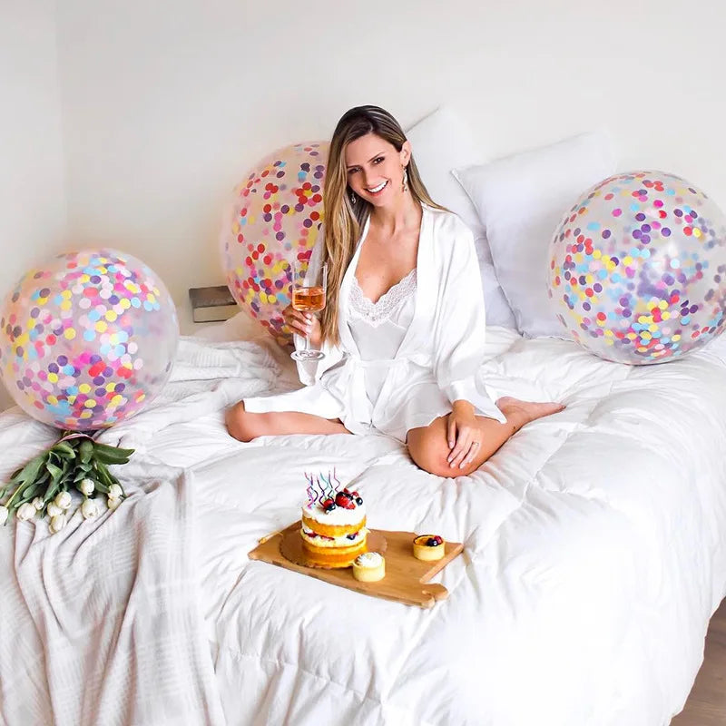 Woman in white robe sits on a bed with colorful balloons, holding a glass of wine. A small cake and pastries are on a tray. White tulips are beside her, adding elegance.