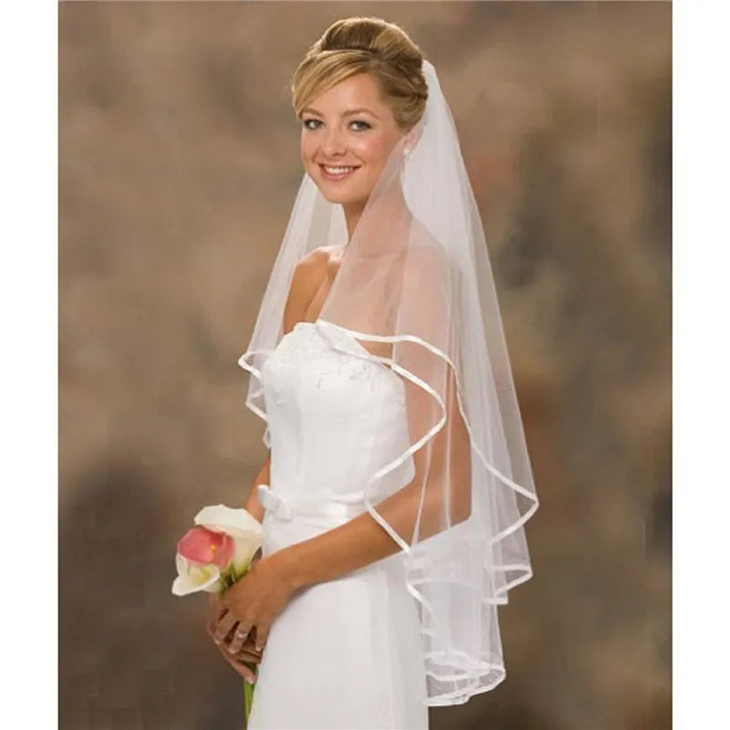 Smiling bride in a white strapless gown with lace details, holding a pink and white calla lily bouquet. She wears a sheer veil over her updo against a soft brown background.