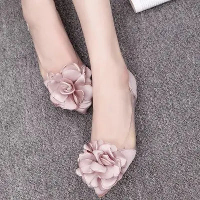 Close-up of feet wearing pink flats adorned with large, decorative fabric flowers on the toe caps, against a gray carpet background.