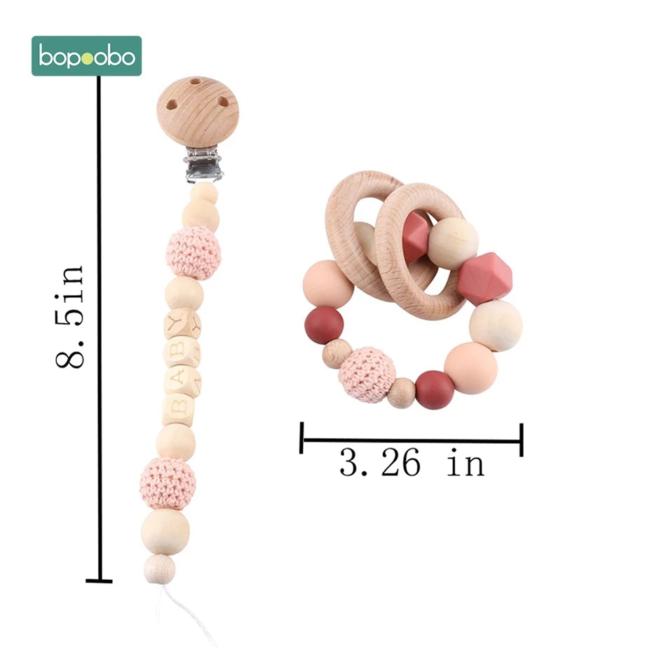 Wooden baby pacifier clip and teething ring set. The clip is 8.5 inches with crochet and bead details, spelling "BABY." The teething ring is 3.26 inches with wooden and silicone beads.