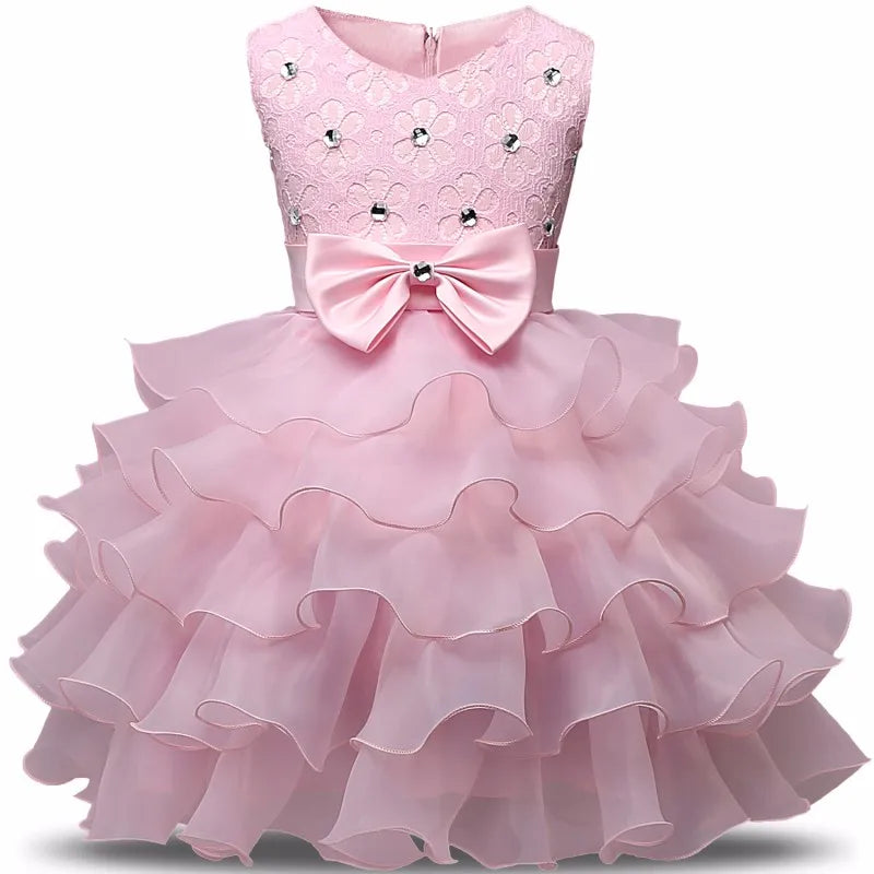 Pink girl's dress with a lace bodice adorned with small flowers and gems, featuring a large bow at the waist. The skirt is layered with cascading ruffles.