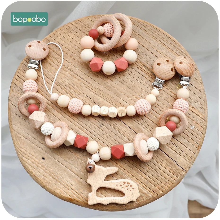 Wooden baby pacifier clip and teether set with beads spelling "BABY," featuring natural wood and pink crochet elements, arranged on a round wooden surface.