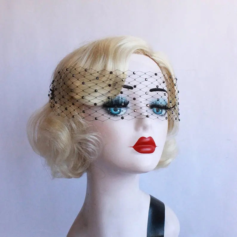 Blonde mannequin head with a black netted veil over blue eyes and red lips, styled in retro fashion. The mannequin wears a black outfit against a neutral background.