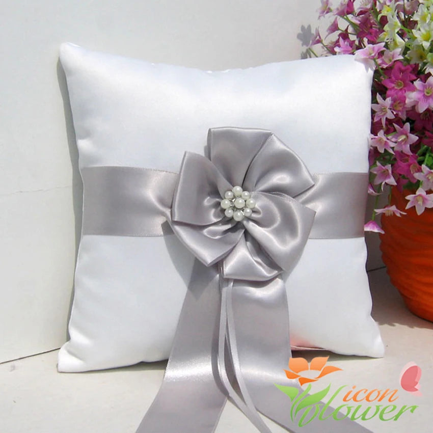 A white satin pillow with a silver ribbon and a decorative flower featuring pearl accents. In the background, a bouquet of pink and white flowers is partially visible.