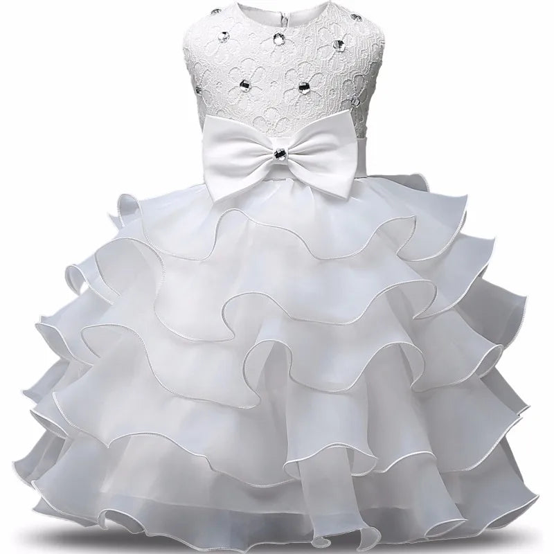 Elegant white baby dress with a lace bodice adorned with gems, a large central bow, and a layered, ruffled skirt. Perfect for special occasions or celebrations.