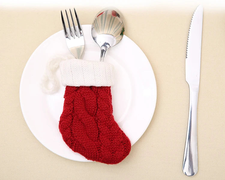 A fork and spoon are tucked into a red knitted Christmas stocking placed on a white plate, with a knife on the right, on a beige tablecloth.
