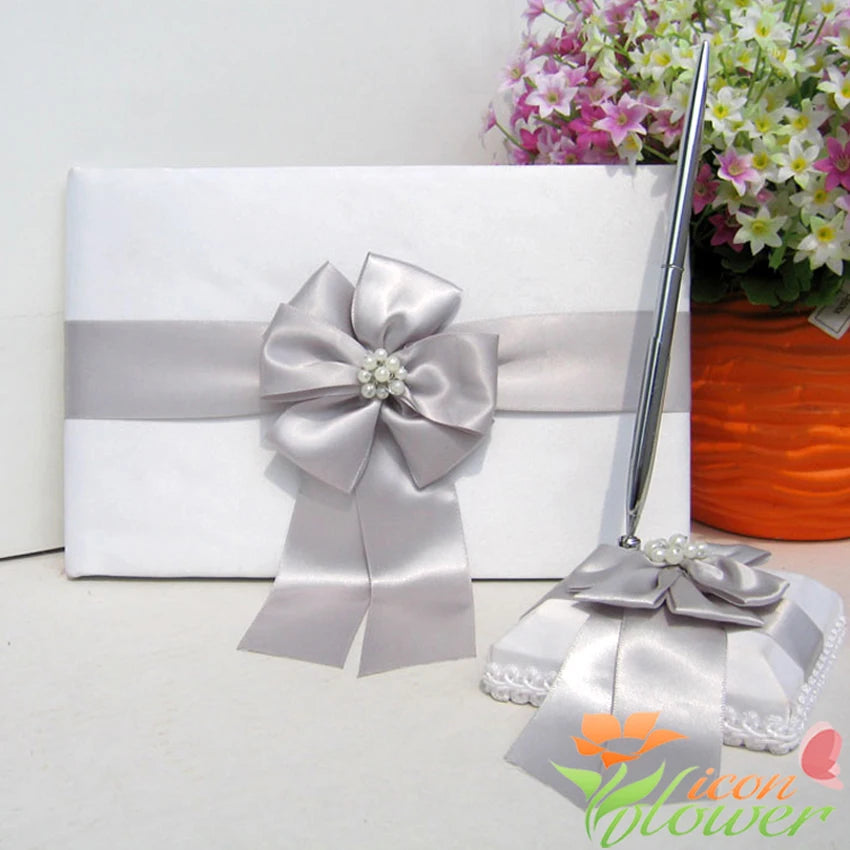 Elegant white guest book with a silver ribbon and pearl-accented bow, accompanied by a matching pen on a padded base, set in front of a bouquet of pink flowers.