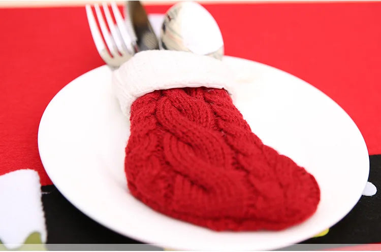 A red knitted Christmas stocking-shaped cutlery holder with a white cuff lies on a white plate, holding a spoon, knife, and fork, all on a red tablecloth.