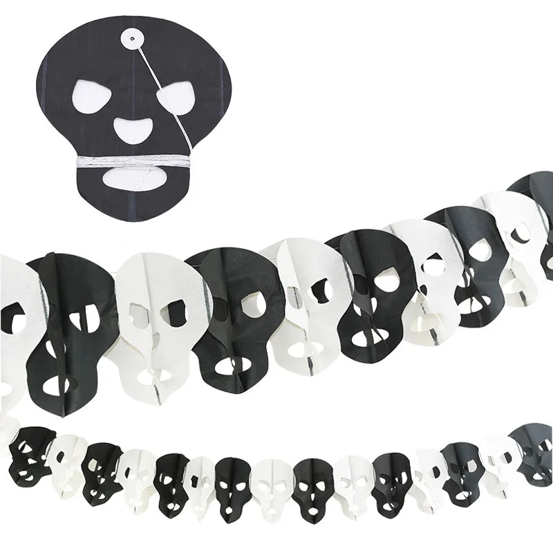 A paper garland features alternating black and white skull shapes. The skulls have hollow eyes and mouths, and they are connected in a chain. Above, a single black skull shows its flat design, with visible folds highlighting how the garland is constructed. The setup conveys a festive, spooky theme.