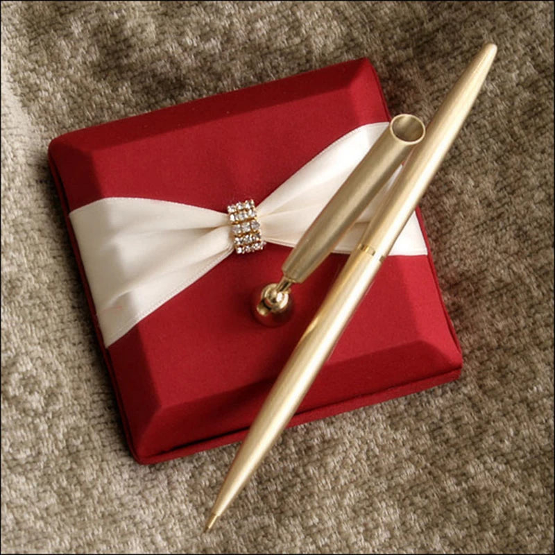 A gold pen rests on a red satin box adorned with a white ribbon and rhinestone detail, placed on a textured beige fabric.