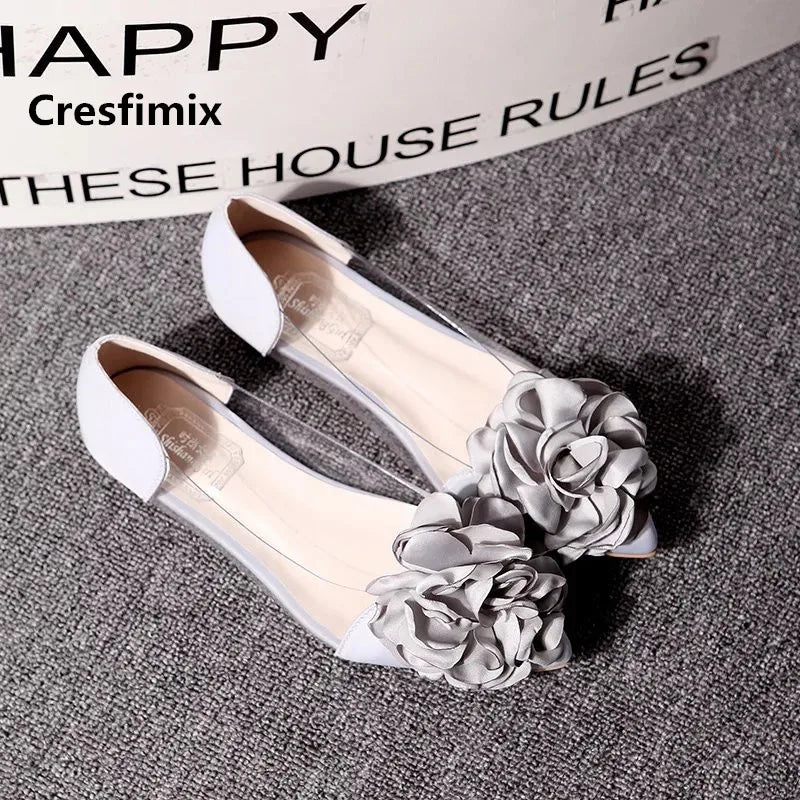 White open-toe flats with large gray floral decorations on a textured gray carpet. Background shows partially visible text.