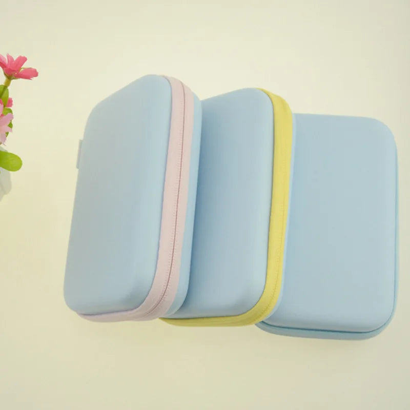 Three light blue rectangular cases with pastel-colored zippers in pink, yellow, and blue, overlapping each other on a light background, next to a small plant with pink flowers.
