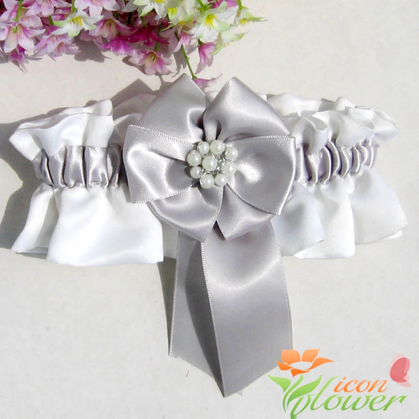 White and silver satin garter with a large bow featuring pearl accents. The background includes pink and white flowers, adding a romantic touch.