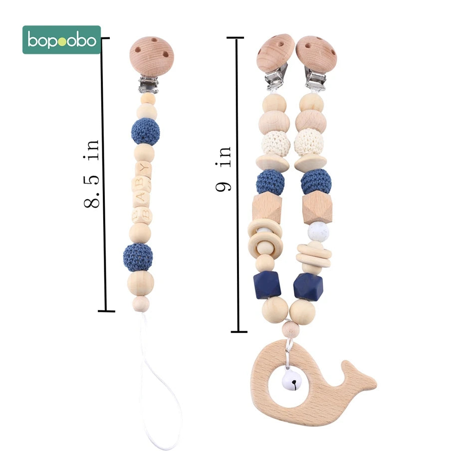 Two pacifier clips made of wooden and crocheted beads in blue and beige tones. One clip features a whale shape with a bell. Lengths are 8.5 and 9 inches.