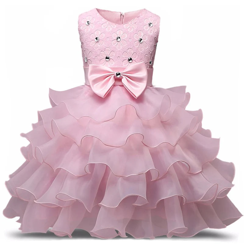 Pink sleeveless dress with lace top, adorned with white and silver floral accents, and a large bow at the waist. The skirt features tiered ruffles for added volume.