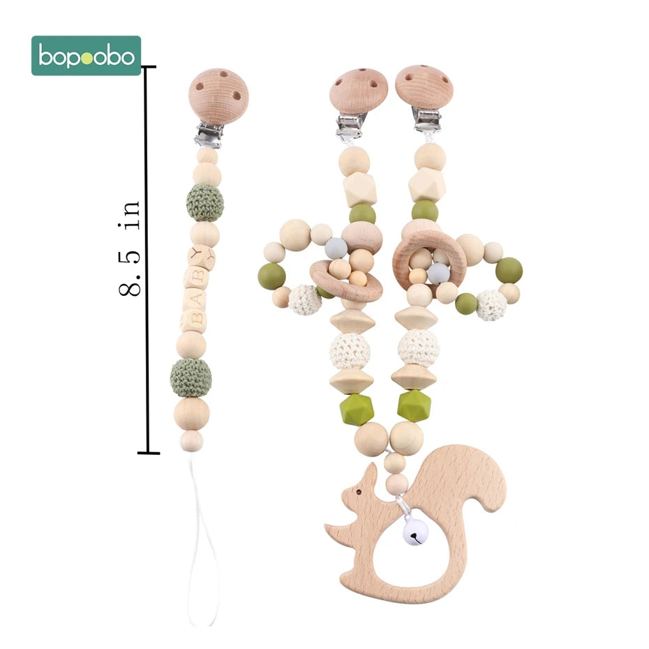 Three pacifier clips featuring wooden beads, crochet details, and green accents. One clip spells "BABY," and another has a wooden squirrel with a small bell. Length is 8.5 inches.