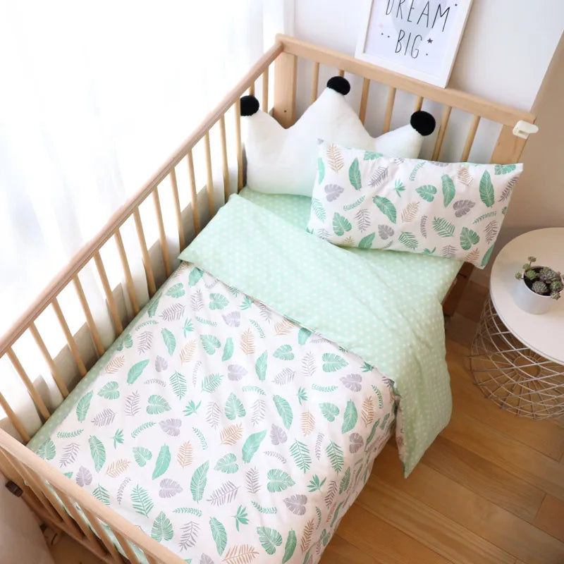 Wooden crib with leaf-patterned bedding and mint green accents. A white cushion with crown, small side table with plant, and "Dream Big" framed art nearby.