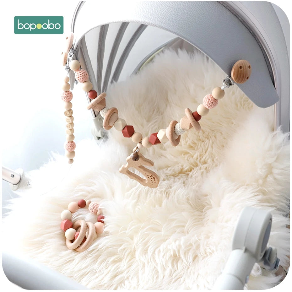 A baby stroller with a fluffy white lining features a hanging wooden and crocheted bead garland, adorned with rings and a small bunny. The brand logo "bopoobo" is visible.